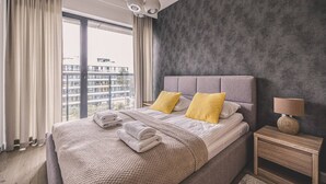 Un dormitorio moderno con una cama grande, una mesita de noche y vistas a un paisaje urbano a través de la ventana.