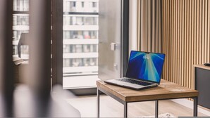 Una computadora portátil sobre una mesa de madera en una habitación moderna con un ventanal de vidrio que da a un balcón.