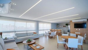 A modern living room with a sofa, dining table, and chairs, a kitchen area, and large windows offering a city view. - Sea View Studio in Condominium With Pool Edf1707 (Salvador)
