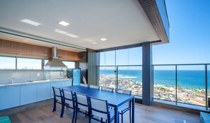 Una cocina moderna con una isla grande, un comedor con mesa y sillas, y un balcón con vistas al océano y al paisaje urbano.