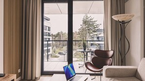 A modern room with a laptop on a table, a chair, and a view of a balcony with buildings and trees. - Puro - Modern Apartment by the Dunes (Trzebiatow)