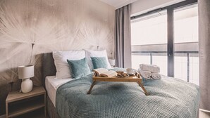 A hotel room with a large bed, a bedside table with a lamp, a tray with breakfast items, and a view of the city through the window.