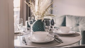 A dining table set with white plates, bowls, and cutlery, a vase with dried flowers, and a green vase with a polka dot pattern.