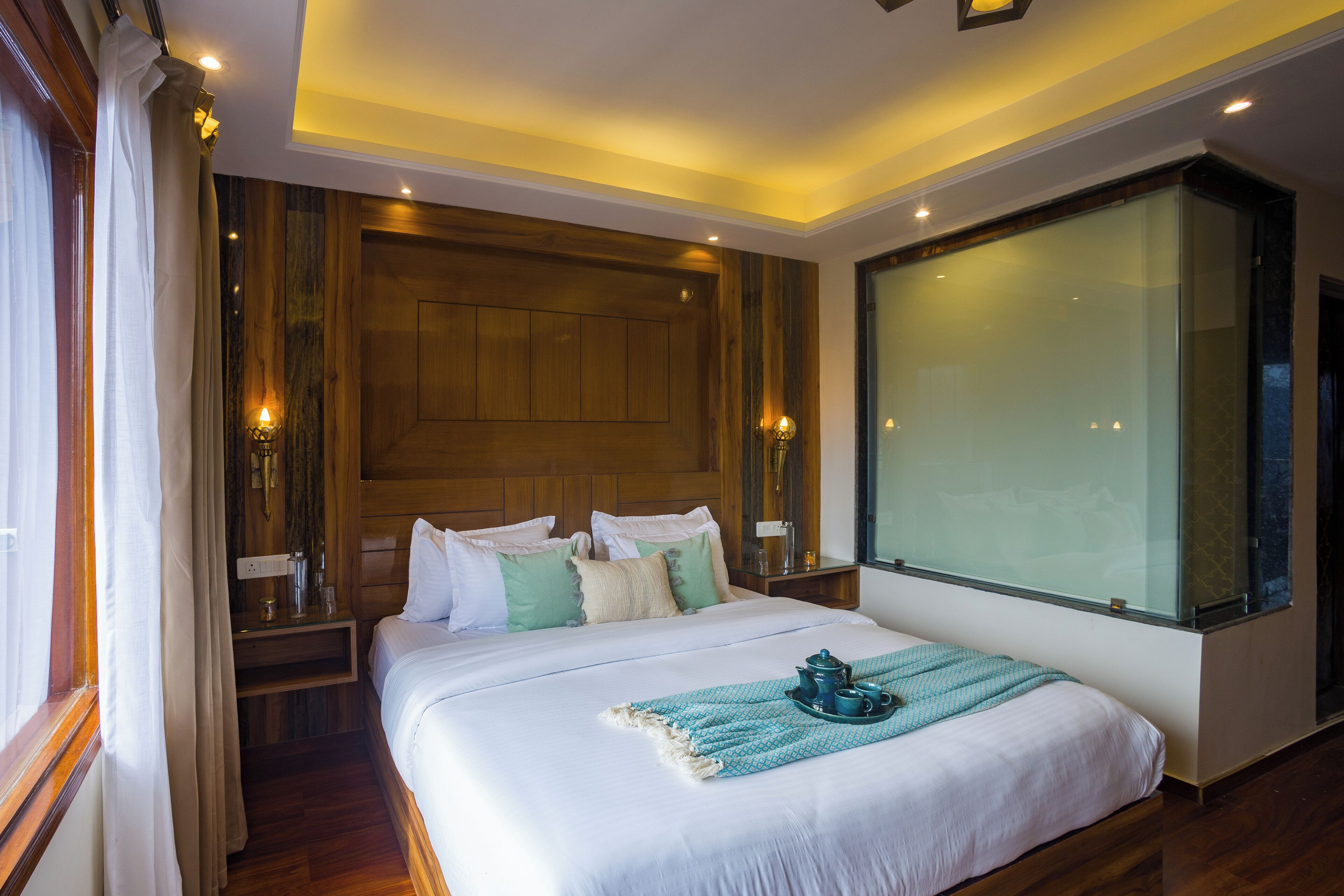 A hotel room with a large bed, wooden headboard, bedside tables with lamps, a window with curtains, and a large mirror.