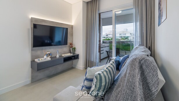 A modern living room with a flat-screen TV, grey sofa, and a sliding glass door leading to a balcony. - Apart in Cond. w/ Pool 400m From the Beach Tdb0101 (Florianópolis)