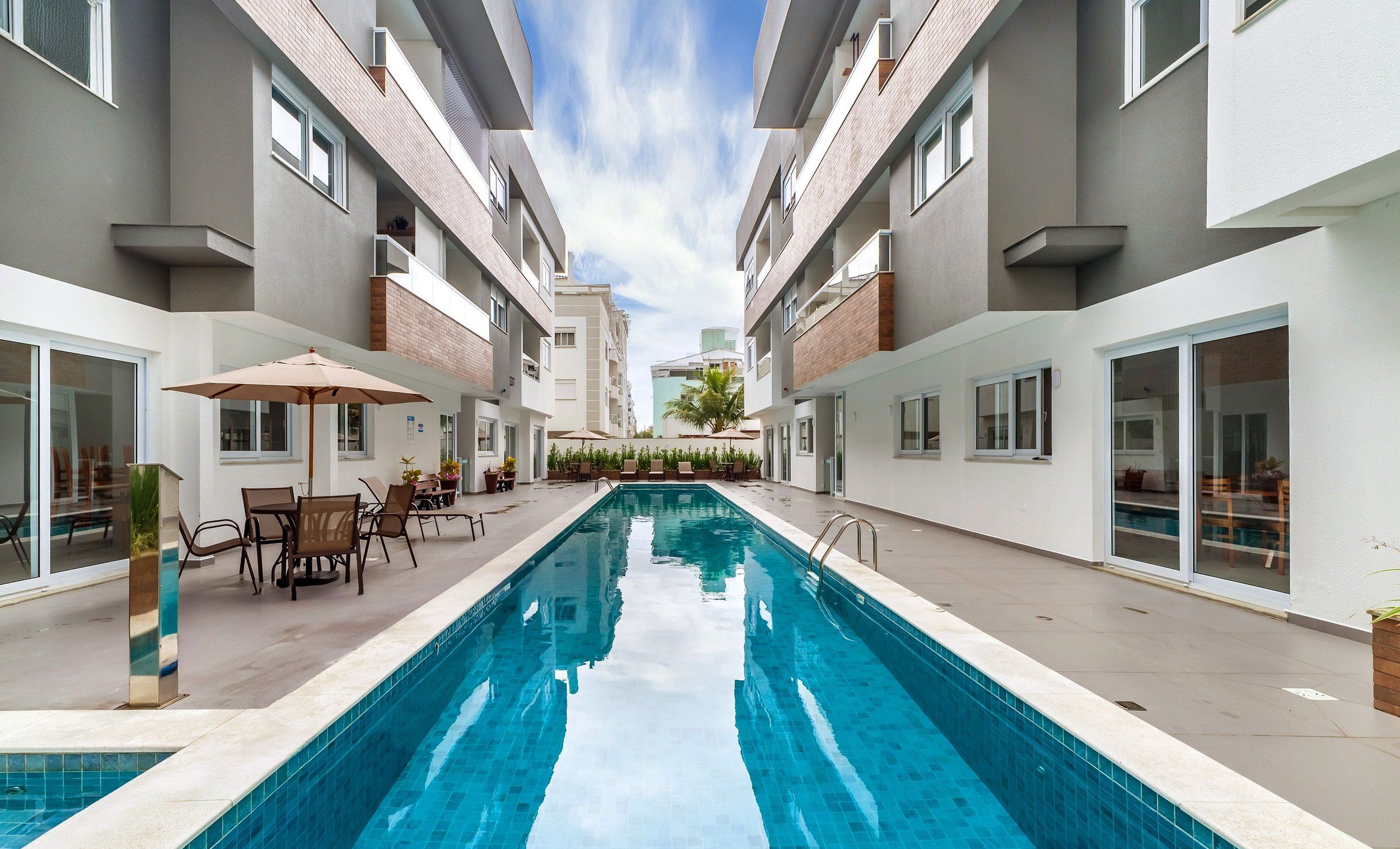 A modern outdoor swimming pool with a clear blue water surface, surrounded by a patio area with chairs and umbrellas, and adjacent multi-story buildings with balconies and large windows.