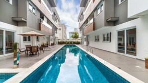 A modern outdoor swimming pool with a clear blue water surface, surrounded by a patio area with chairs and umbrellas, and adjacent multi-story buildings with balconies and large windows. - Apart in Cond. w/ Pool 400m From the Beach Tdb0101 (Florianópolis)
