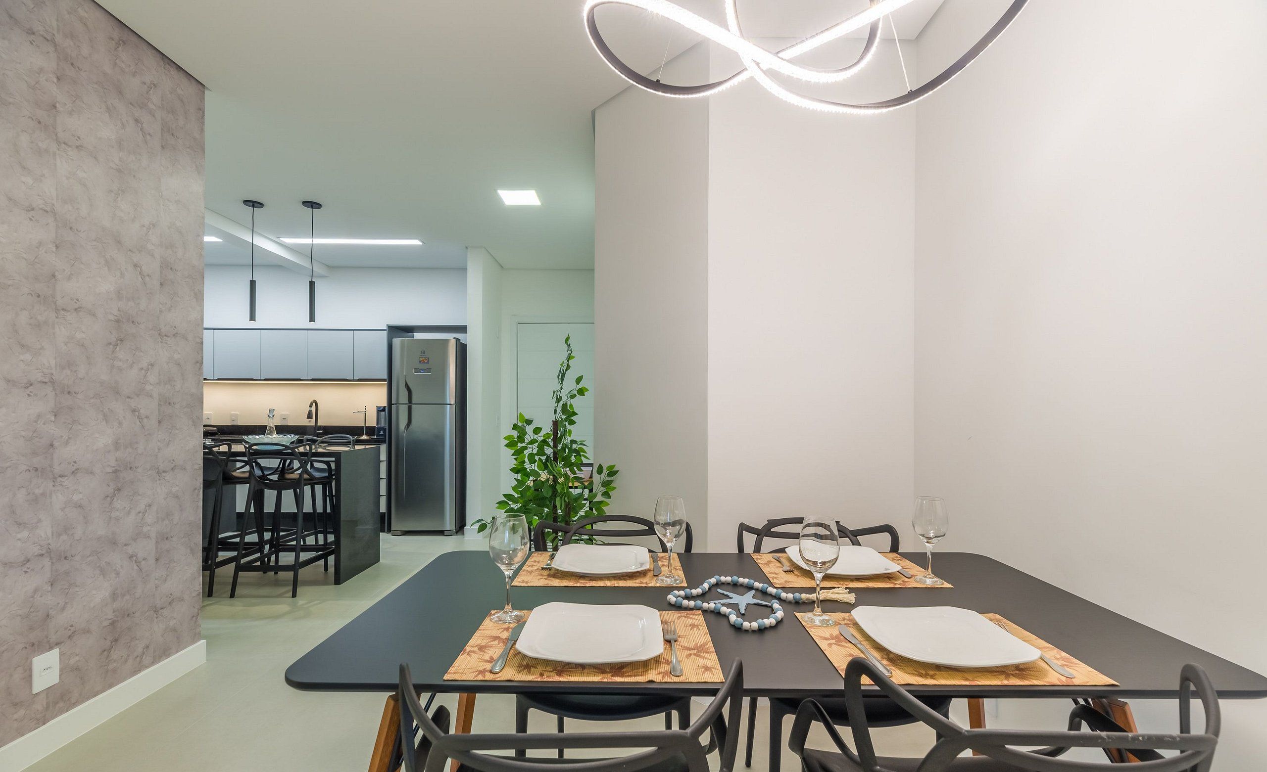 Un moderno comedor con una mesa negra dispuesta para cuatro personas, rodeada de sillas negras. Se ve una planta al fondo, y la habitación tiene un diseño minimalista con paredes blancas y piso gris.
