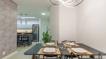 Un moderno comedor con una mesa negra dispuesta para cuatro personas, rodeada de sillas negras. Se ve una planta al fondo, y la habitación tiene un diseño minimalista con paredes blancas y piso gris.