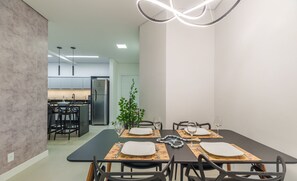 Un espace repas moderne avec une table noire dressée pour quatre personnes, entourée de chaises noires. On aperçoit une plante à l’arrière-plan, et la pièce présente un design minimaliste avec des murs blancs et un sol gris.