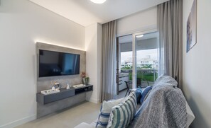 A modern living room with a flat-screen TV, grey sofa, and a sliding glass door leading to a balcony. - Apart in Cond. w/ Pool 400m From the Beach Tdb0101 (Florianópolis)