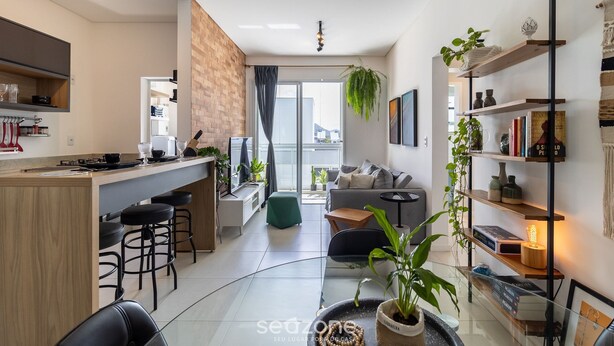 A modern apartment with a kitchen, dining area, and living room. It features a bar area with stools, a sofa, and a coffee table. There are plants and books on shelves, and a large window providing natural light. - CDN - Apartments in Campeche (Florianópolis)