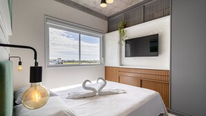 A hotel room with a bed, a mounted TV, a window with a view, and a wall-mounted light fixture. - CDN - Apartments in Campeche (Florianópolis)