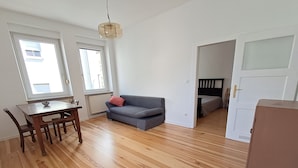 Een kamer met een houten vloer, een grijze bank, een eettafel met stoelen en een witte deur.