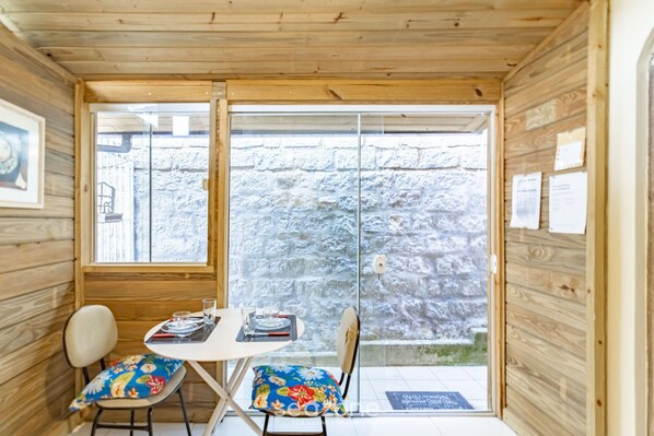 Una pequeña habitación de madera con una mesa, sillas, un ventanal y una pared de piedra en el exterior.