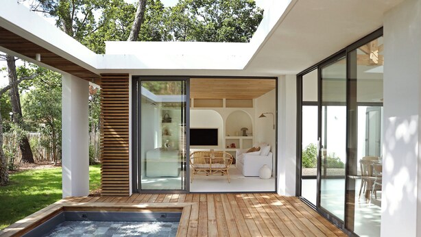 A modern house with a wooden deck, a hot tub, and a glass door leading to a living area with a sofa and TV.