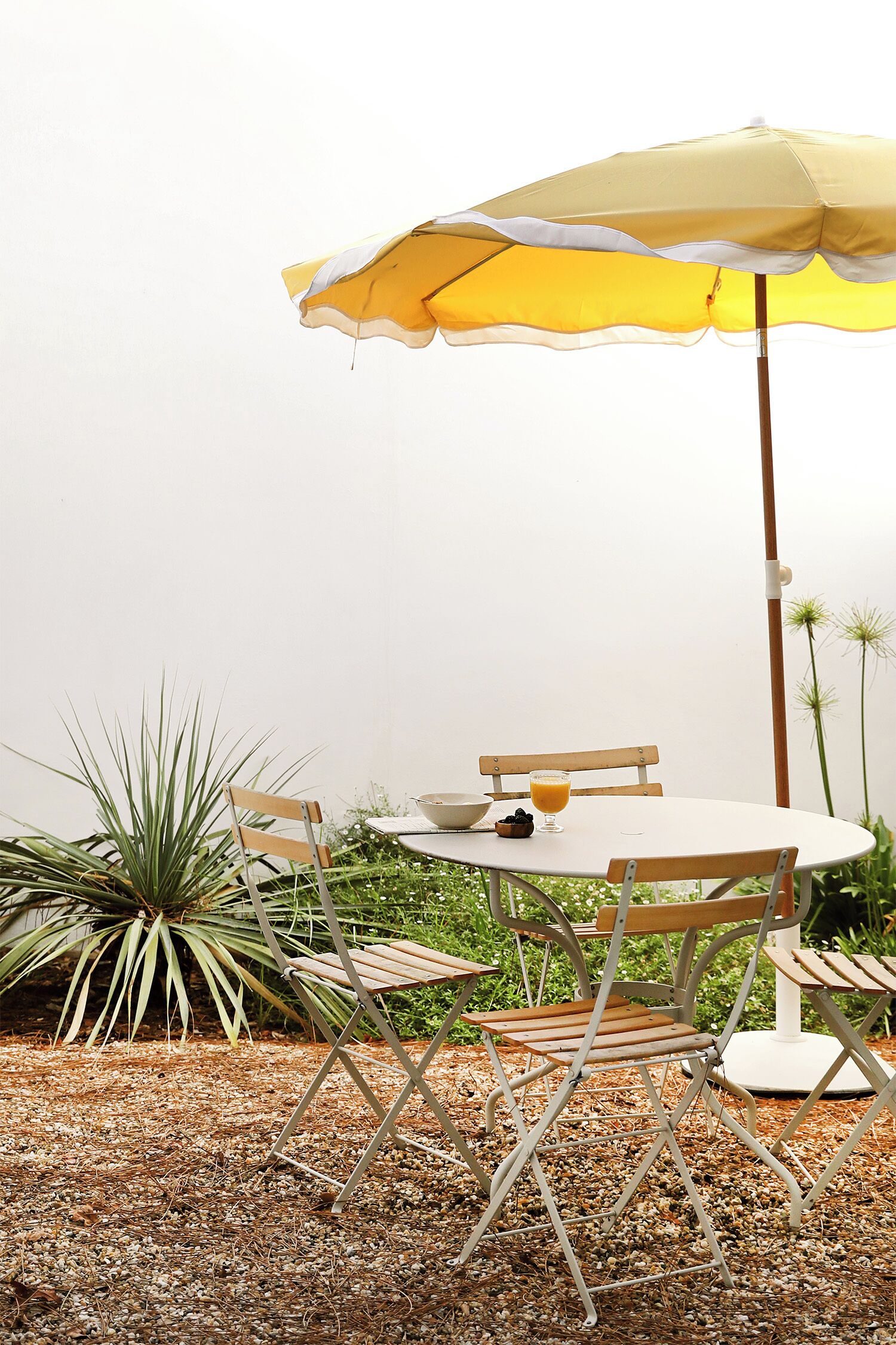 A round table with a yellow umbrella, surrounded by wooden chairs, set on a gravel surface with plants in the background.