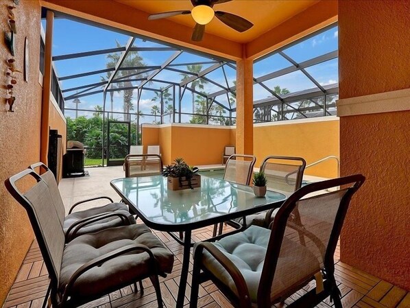 A glass-enclosed patio with a table and chairs, a ceiling fan, and a view of palm trees. - Adorable Townhome Remodeled With Comfort And Relaxation In Mind! Splash Pool! 4 Bedroom Townhouse (Kissimmee)