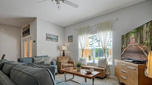 Un salon comprenant un canapé, des fauteuils, une table basse et un téléviseur. On y trouve également des rideaux, une lampe et un tableau accroché au mur.