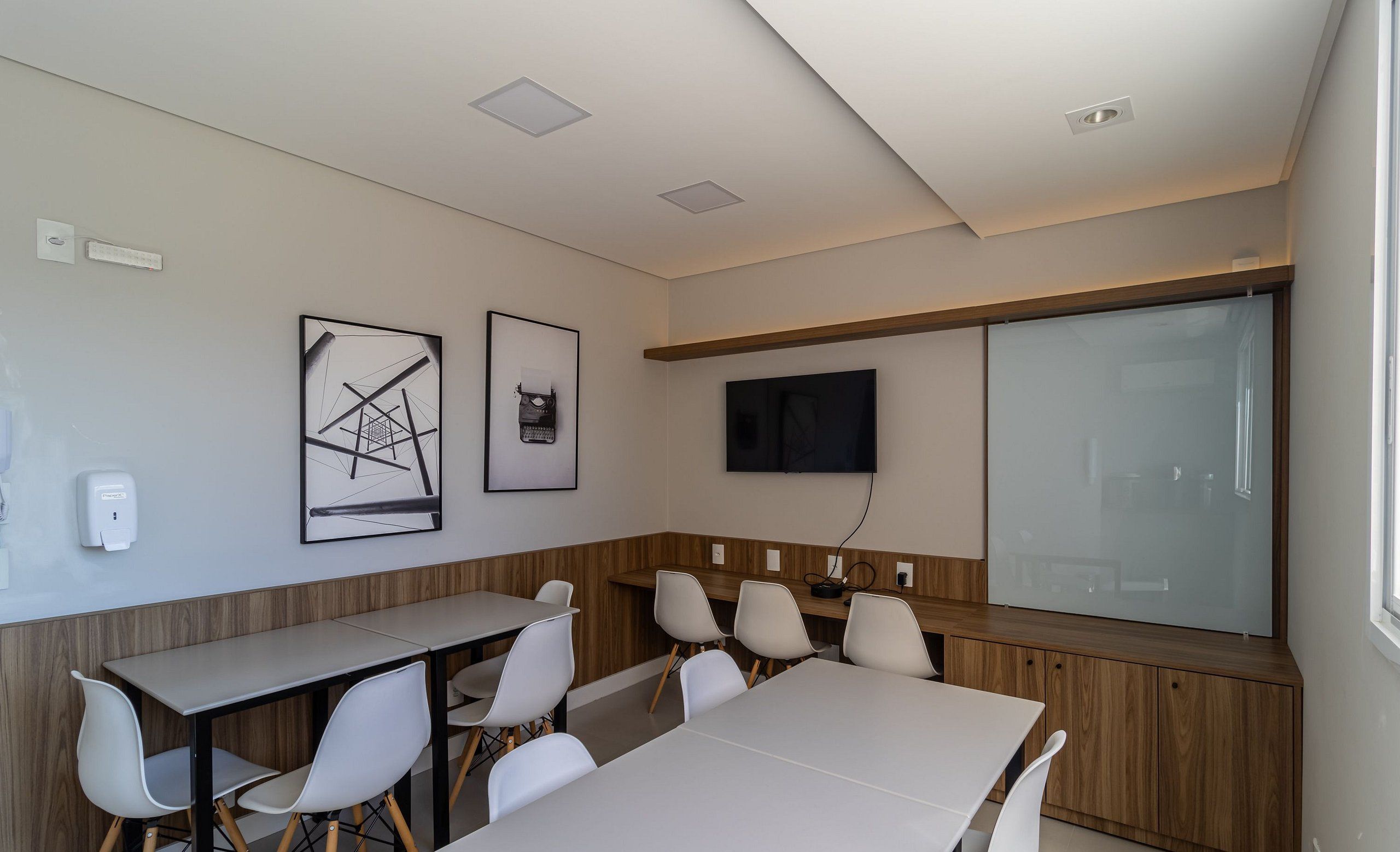 Un salón de conferencias con una larga mesa, sillas blancas, un televisor montado y obras de arte enmarcadas en la pared.