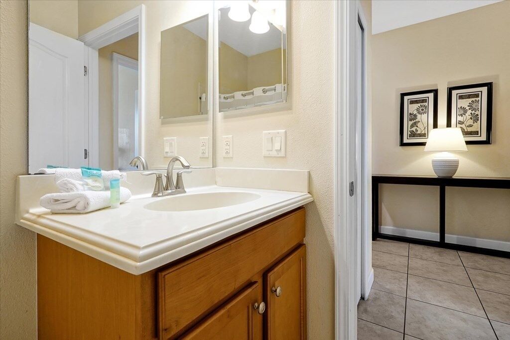A bathroom with a double sink, wooden vanity, and a mirror. There are framed pictures on the wall and a lamp on a side table.