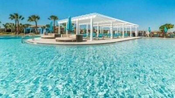 A clear swimming pool with a covered seating area and palm trees in the background. - Magical Getaway Near Disney Private Pool + Resort Access, Bbq Grill! 4 Bedroom Townhouse (Kissimmee)