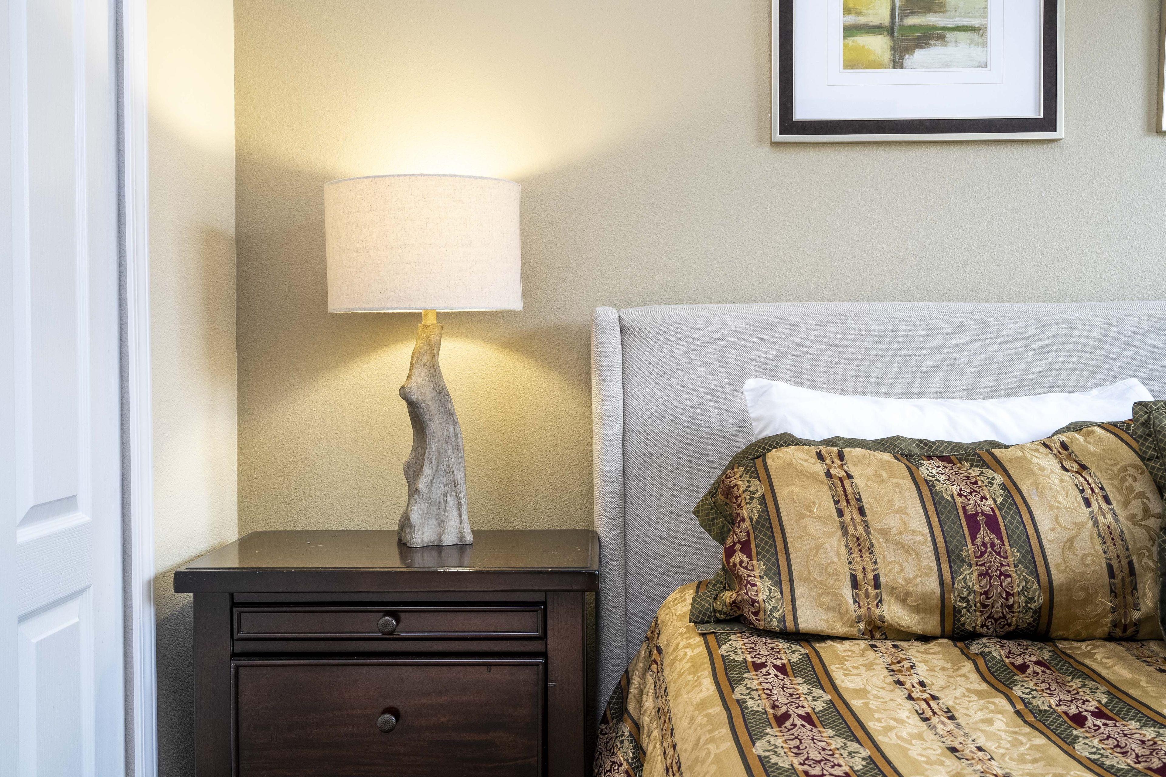 Un lit bien fait, avec une literie à motifs et un seul oreiller, une table de chevet avec une lampe et un tableau encadré accroché au mur.