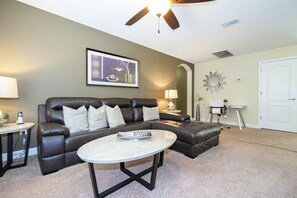 A living room with a leather sofa, a coffee table, a lamp, and a ceiling fan. - Family Pool Home Near Disney!! Modern Decor, Bbq ,resort W/ Tons Of Amenities! 6 Bedroom Home (Kissimmee)