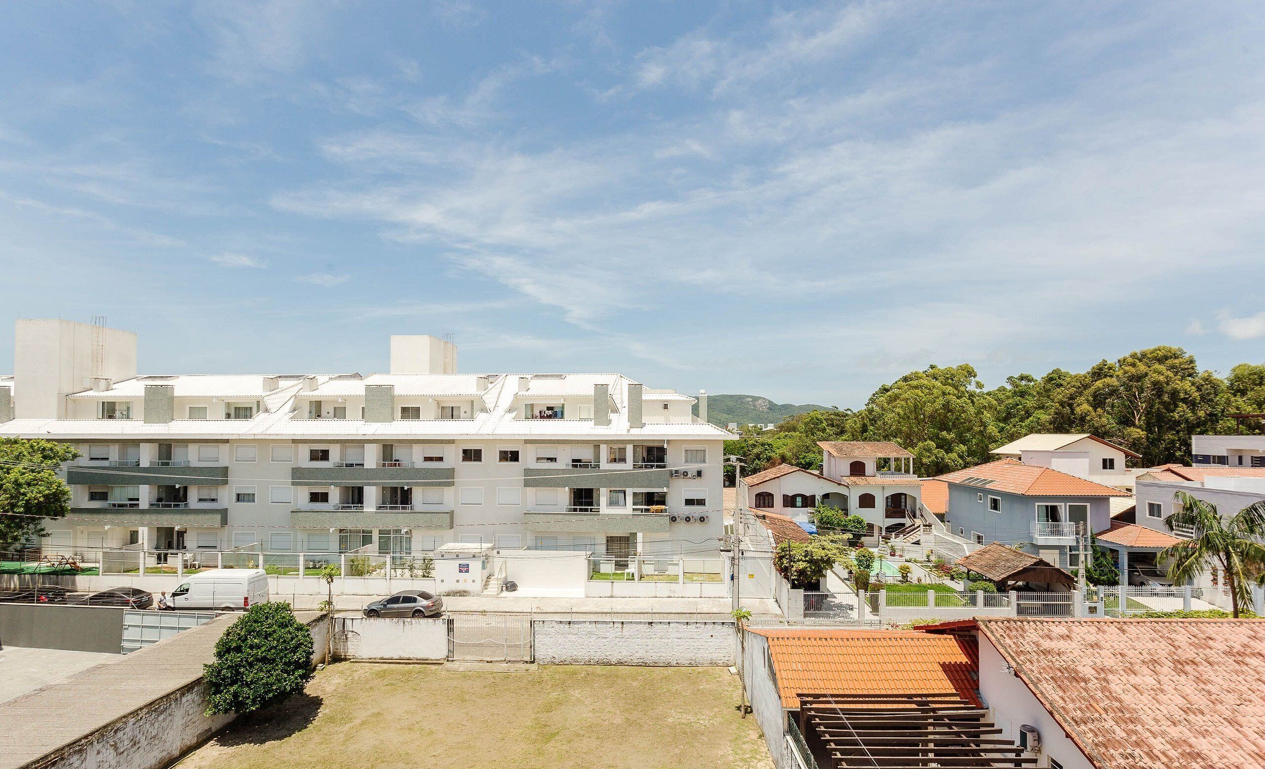 A multi-story residential building with balconies, a parking area, and a view of a hilly landscape.