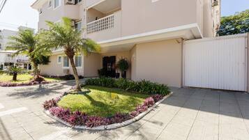 A multi-story residential building with a gated entrance, a small garden with palm trees, and a tiled pavement.
