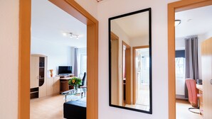 A modern living room with a flat-screen TV, a glass coffee table, and a sofa. There is a large mirror on the wall and a wooden door leading to another room. - acora Bremen Garden Living the City (Bremen)