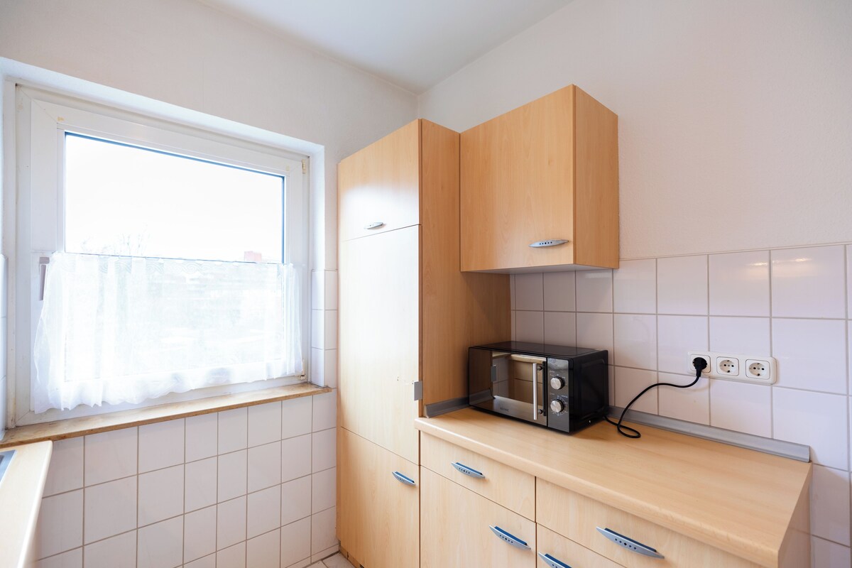 A kitchen with wooden cabinets, a microwave, and a window with a curtain.