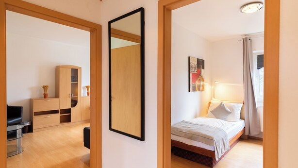 A bedroom with a bed, bedside table, and a mirror. There is a wooden wardrobe and a desk with a chair in the adjacent room. - acora Bremen Garden Living the City (Bremen)