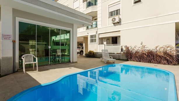 A swimming pool with a glass door entrance, white chairs, and a building with balconies. - Apto espaçoso com piscina 750m da praia | CHR025 (Florianópolis)
