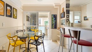A modern kitchen with a glass dining table and colorful chairs, a bar area with stools, and a view of the outside. - Apto espaçoso com piscina 750m da praia | CHR025 (Florianópolis)