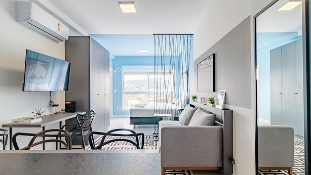 A modern living room with a sofa, a TV, a dining area with chairs, and a large window with a view. - Apartments Next to Ufsc - SVE (Florianópolis)