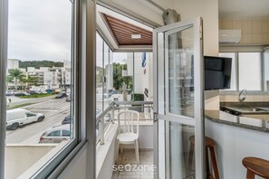 A balcony with a view of a street and buildings, a white chair, and a kitchen area with a TV and sink. - Fully Equipped Apartments Just Minutes From Jurere Beach - CIE (Florianópolis)
