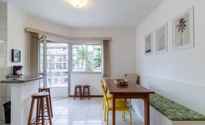 A modern kitchen with a dining area, a bench, and a window with a view of buildings and palm trees. - Fully Equipped Apartments Just Minutes From Jurere Beach - CIE (Florianópolis)