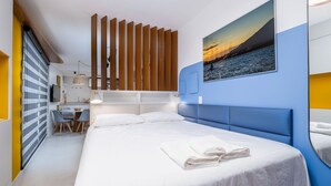 Una habitación de hotel moderna con una cama grande, un escritorio con sillas, una partición de listones de madera y un cuadro de un atardecer sobre el agua.