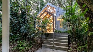 A modern, glass-walled structure with a staircase leading to a wooden deck, surrounded by dense tropical foliage. - Flat Vista mar Dentro de Resort de Luxo Sto2206 (Florianópolis)