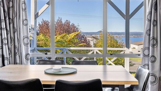 A dining area with a table and chairs, a view of a balcony with plants and a distant landscape.