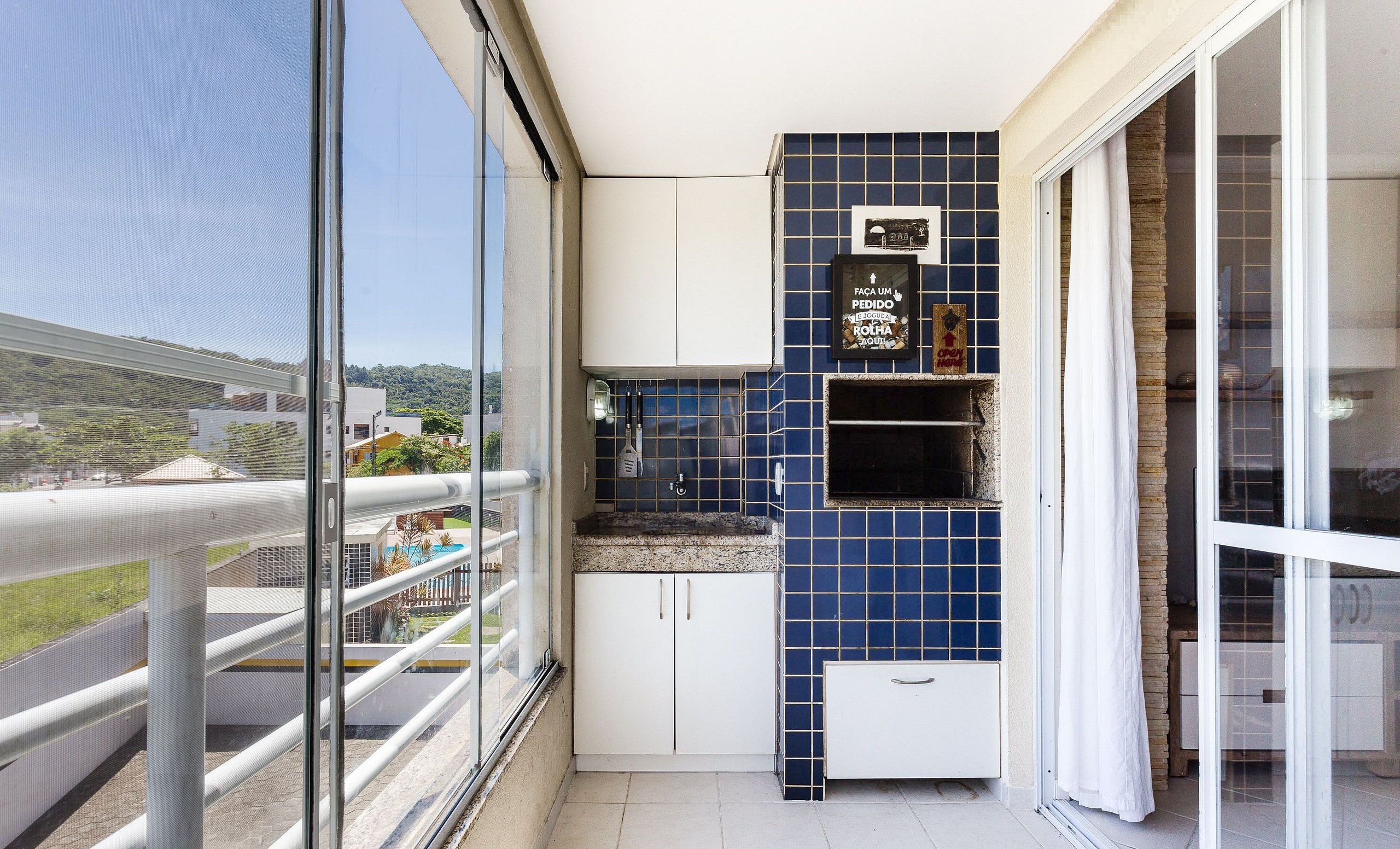 A balcony with a glass railing, a small kitchen area with blue tiles, and a view of a residential area.