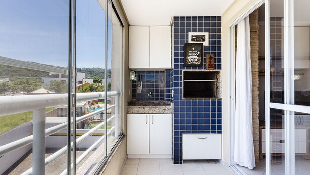 Un balcón con barandilla de vidrio, un pequeño rincón de cocina con azulejos azules y vistas a una zona residencial.