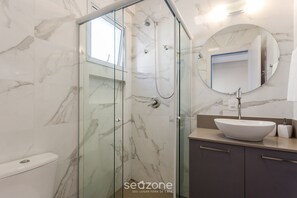 A modern bathroom with a glass-enclosed shower, a round mirror, a white sink, and dark-toned cabinets. - Building With Swimming Pool in Ingleses Norte - STN (Florianópolis)