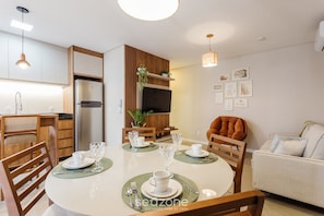 A modern dining area with a round table set for four, wooden chairs, and a kitchen in the background. - Building With Swimming Pool in Ingleses Norte - STN (Florianópolis)