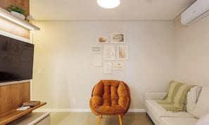 A living room with a flat-screen TV, a brown armchair, a white sofa, and a wall with framed artwork. - Building With Swimming Pool in Ingleses Norte - STN (Florianópolis)