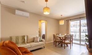 A modern living room with a sofa, dining table, and chairs. There is a large window with a view of buildings. - Building With Swimming Pool in Ingleses Norte - STN (Florianópolis)