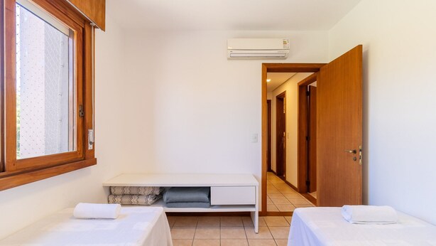A hotel room with two beds, a small table, a window with blinds, an air conditioning unit, and a hallway leading to other rooms. - Charm and Comfort 450m From the Beach! Cmh0202 (Florianópolis)