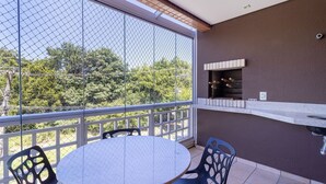 A balcony with a round table and chairs, a brick barbecue grill, and a view of trees through a chain-link fence.