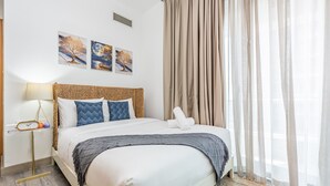 Un lit bien fait, avec une tête de lit en osier, une table de chevet avec une lampe, et trois tableaux encadrés accrochés au mur.